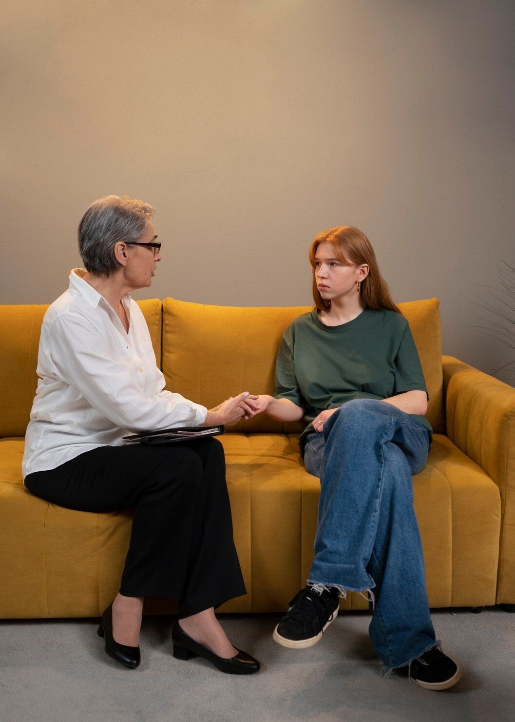 Two people sit on a mustard couch, holding hands and talking in a calm setting.