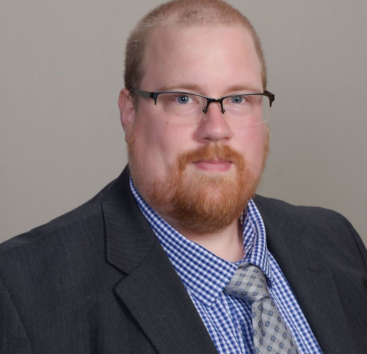 Headshot of a man in glasses, dark blazer, blue checkered shirt, and gray tie on a neutral background