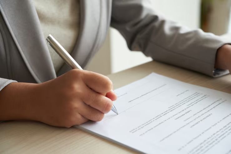 Person in a gray suit writes on a document at a desk.