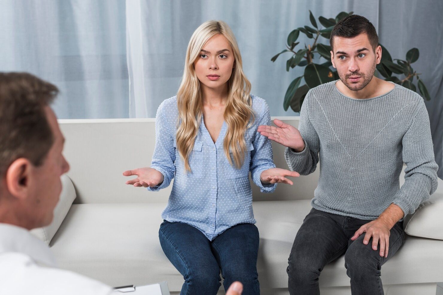 Two people in casual clothes sit on a couch, gesturing as they talk during a conversation with another person.