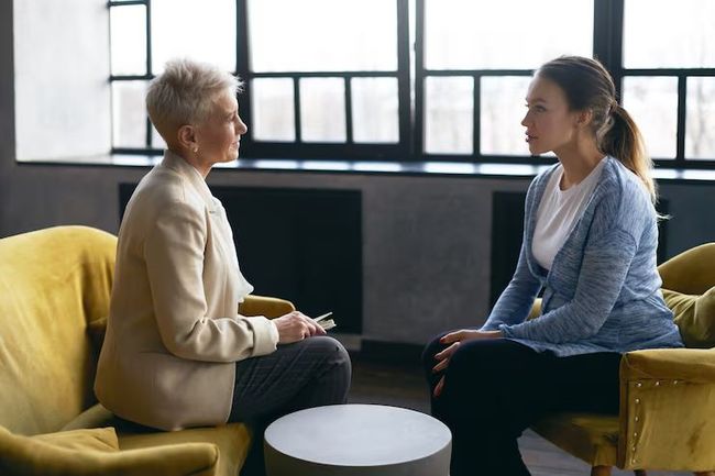 Two people talking on yellow chairs in a bright room with large windows