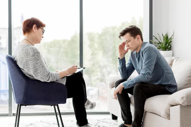 Two people in a bright living room, one taking notes while the other sits with head in hand.