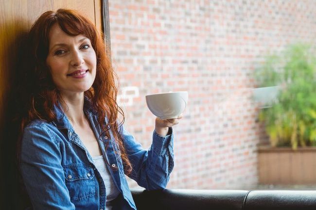 Woman in a denim shirt smiles while holding a white cup in a cozy indoor setting.