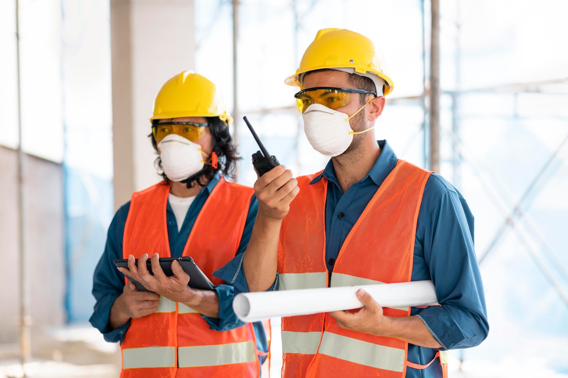 Dois trabalhadores da construção civil usando máscaras e coletes de segurança estão de pé um ao lado do outro.