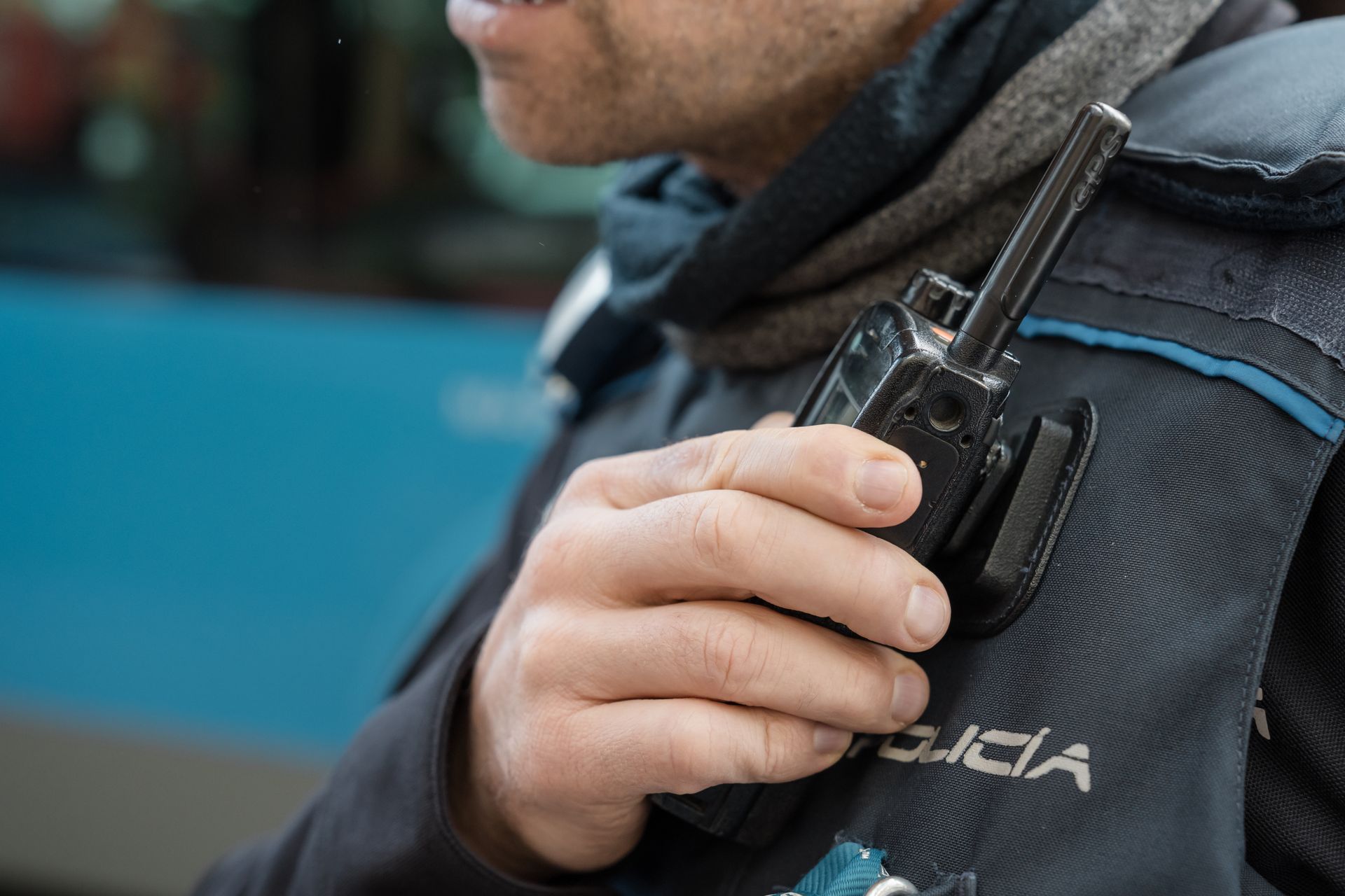 Person holding a handheld radio on a dark uniform beside a blue vehicle.