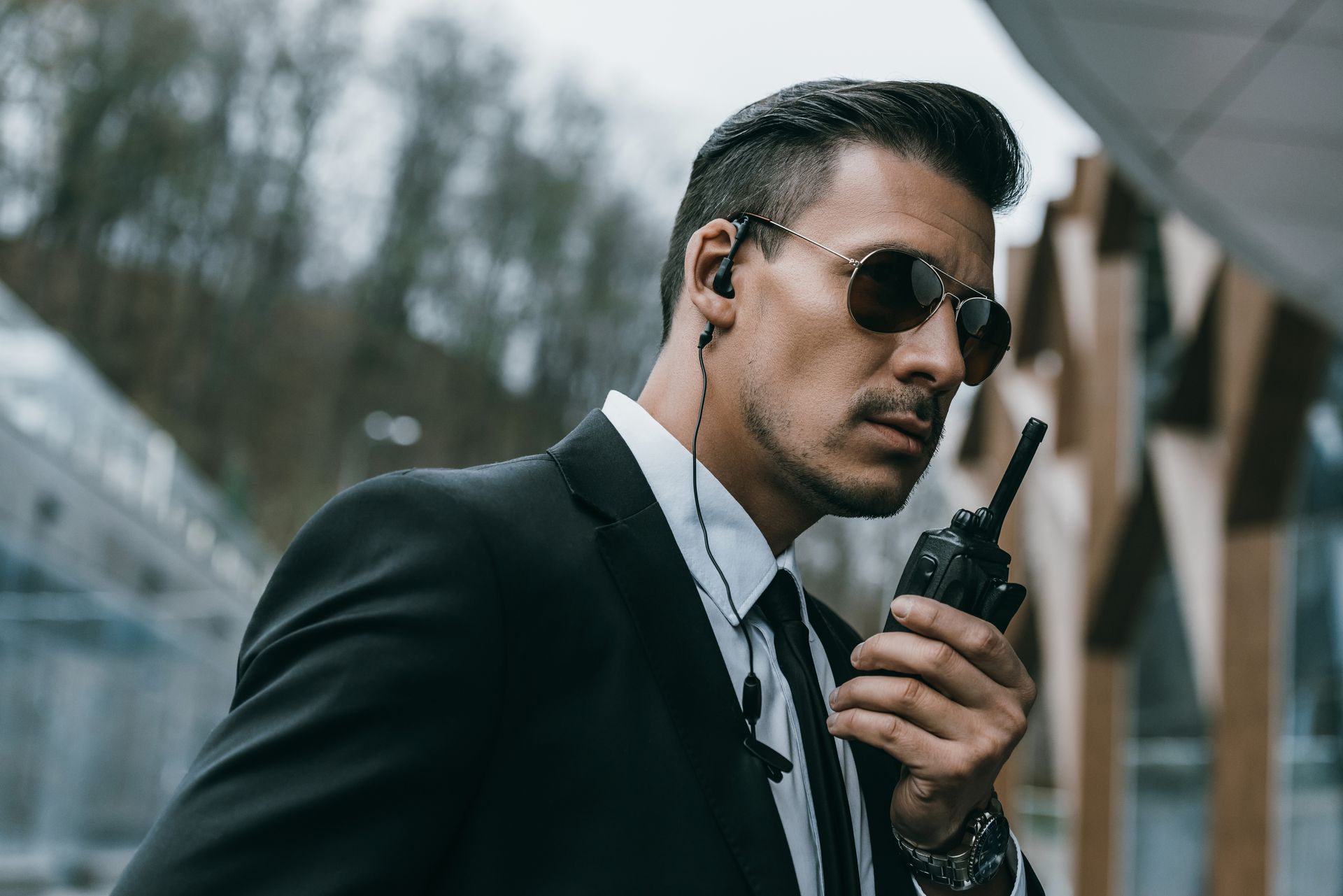Man in a black suit and sunglasses holding a walkie-talkie outdoors near a bridge.