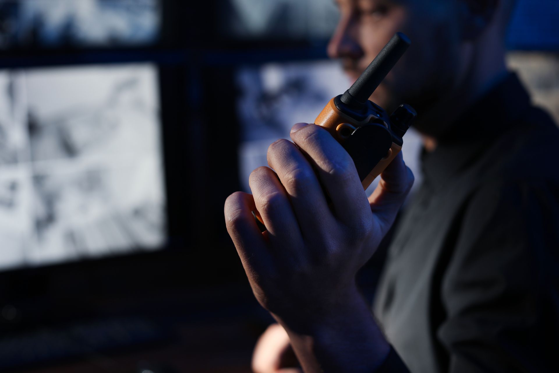 Person holding a handheld radio in a dim control room with monitors in the background.