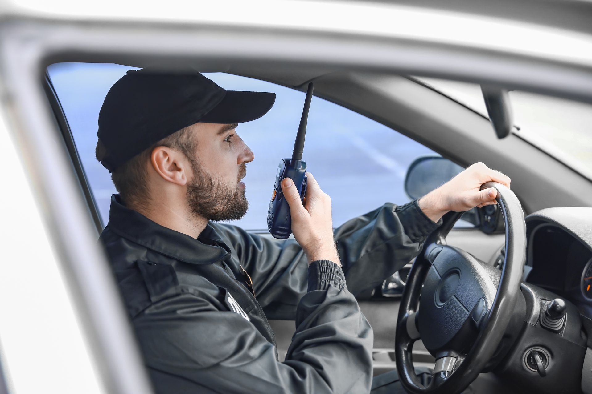 Driver speaking into a walkie-talkie while steering a car, wearing a black cap and jacket.