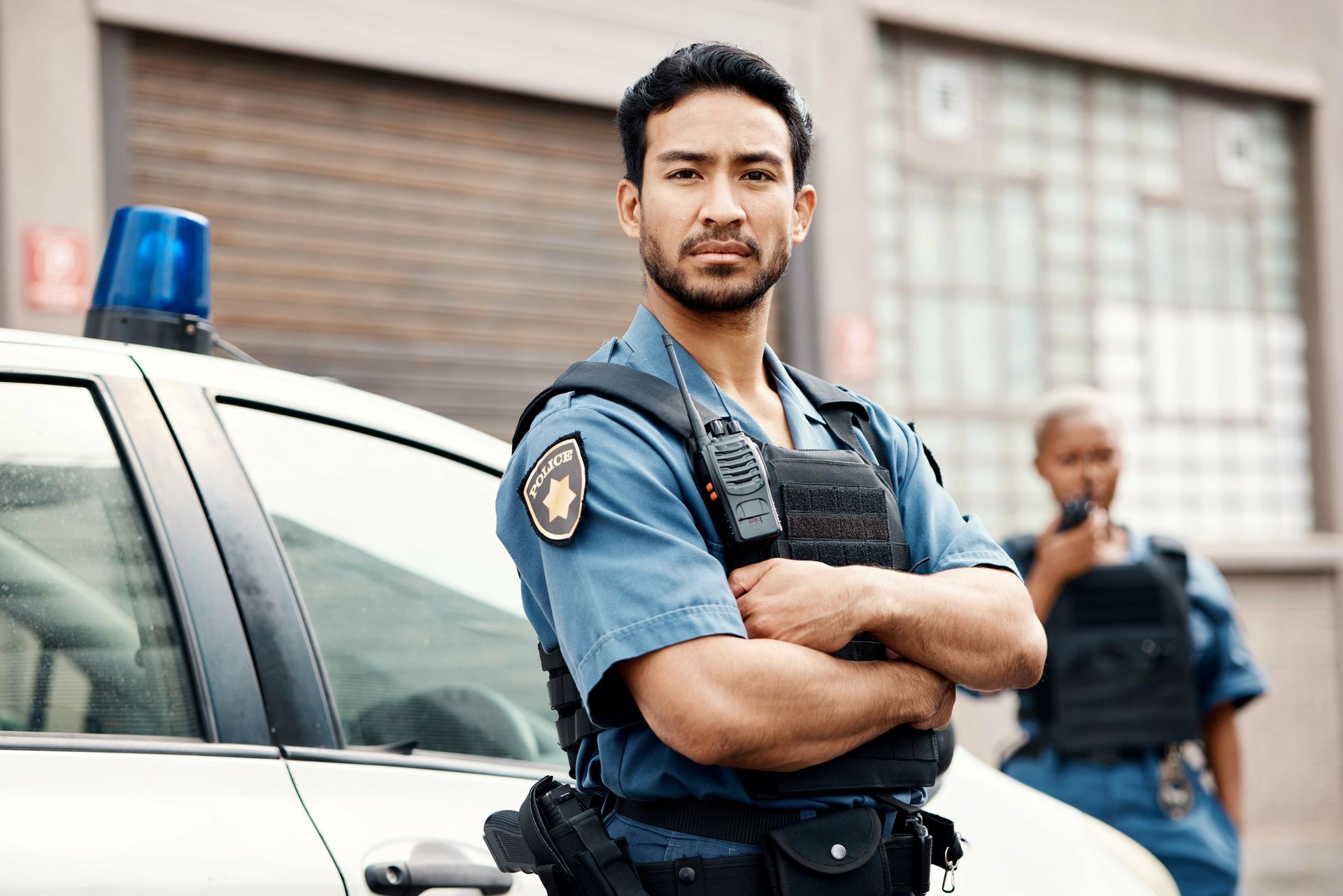Police officer standing beside patrol car with arms crossed, another officer blurred in the background.