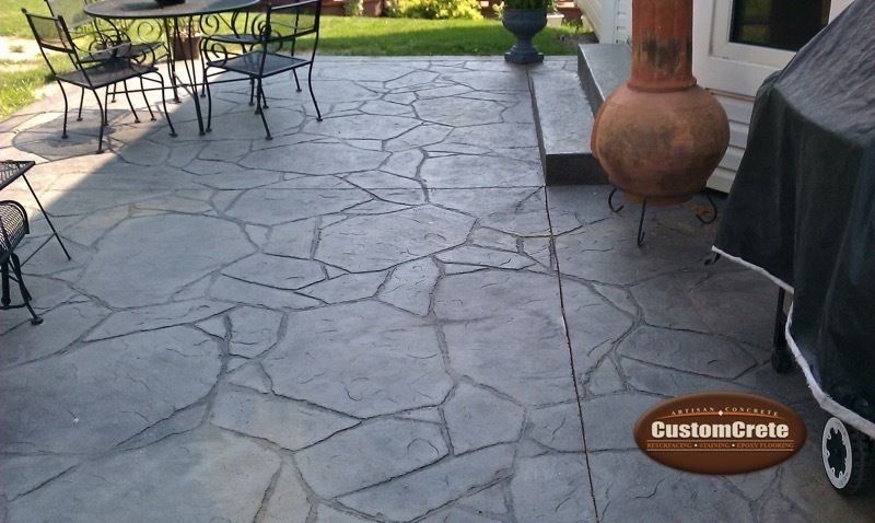 Stone-patterned patio with outdoor table and chairs, potted urn, and covered grill near a house door.