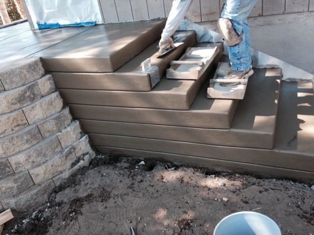 Worker laying brown pavers on outdoor steps beside a stone wall and dirt ground