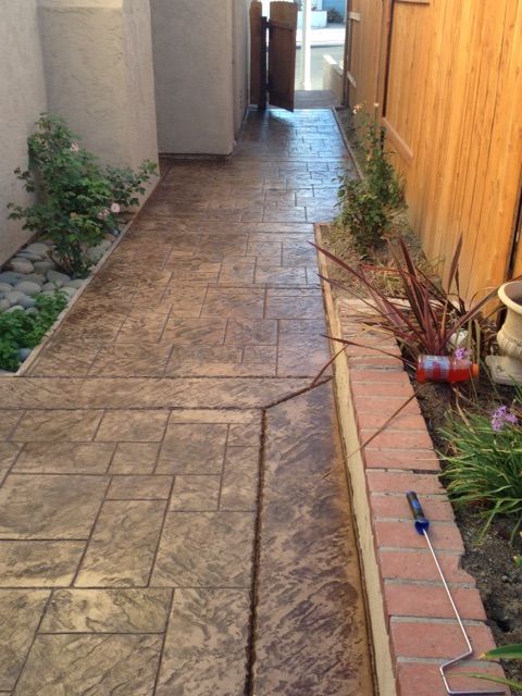 Narrow outdoor walkway with stamped concrete, potted plants, and a wooden fence along the side.