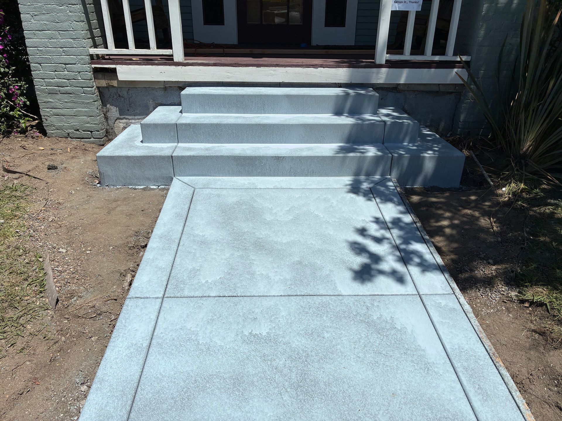 Concrete walkway leading to front steps of a house, with short railings and shadows on the path