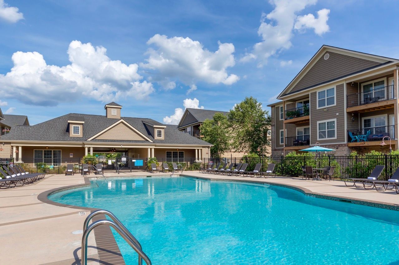 A resort-style swimming pool at an apartment complex, with lounge chairs on the deck and multi-story buildings nearby.