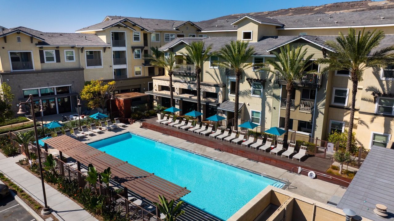 An elevated view of an apartment complex pool area with palm trees, lounge chairs, and teal umbrellas.
