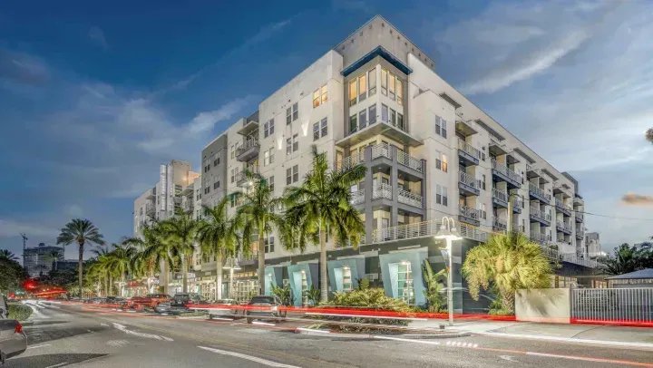 A multi-story modern apartment building with balconies and ground-floor storefronts, lined with palm trees at dusk.