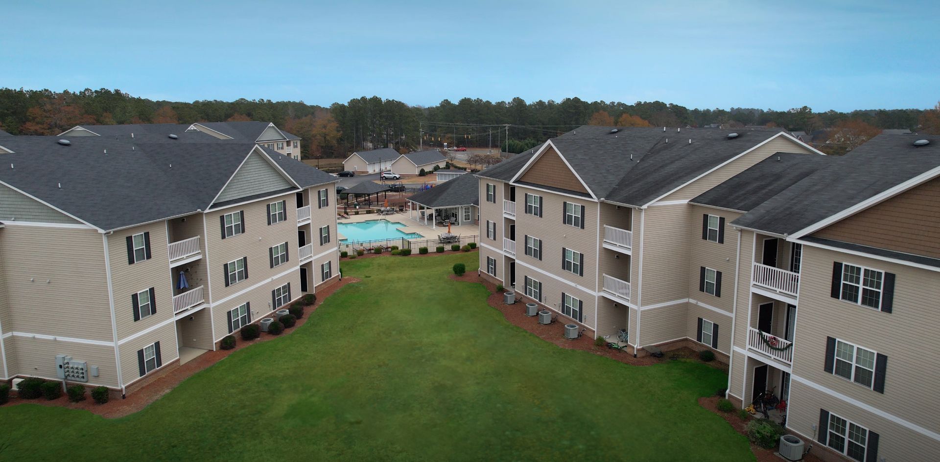 An aerial view of two beige apartment buildings with dark shingled roofs, flanking a central lawn and swimming pool.