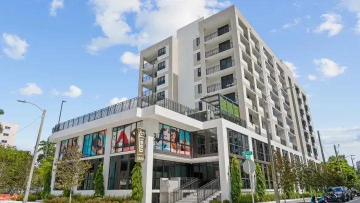 A modern, multi-story building with a white exterior, large ground-floor windows, and balconies under a blue, cloudy sky.