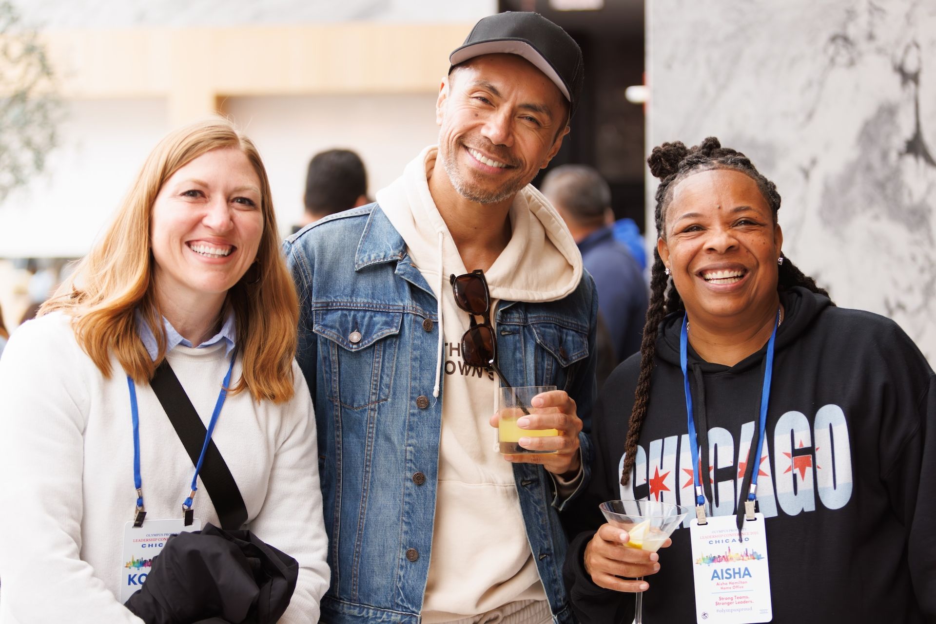 Three people smiling, wearing conference badges and holding drinks in an indoor, neutrally decorated lobby.