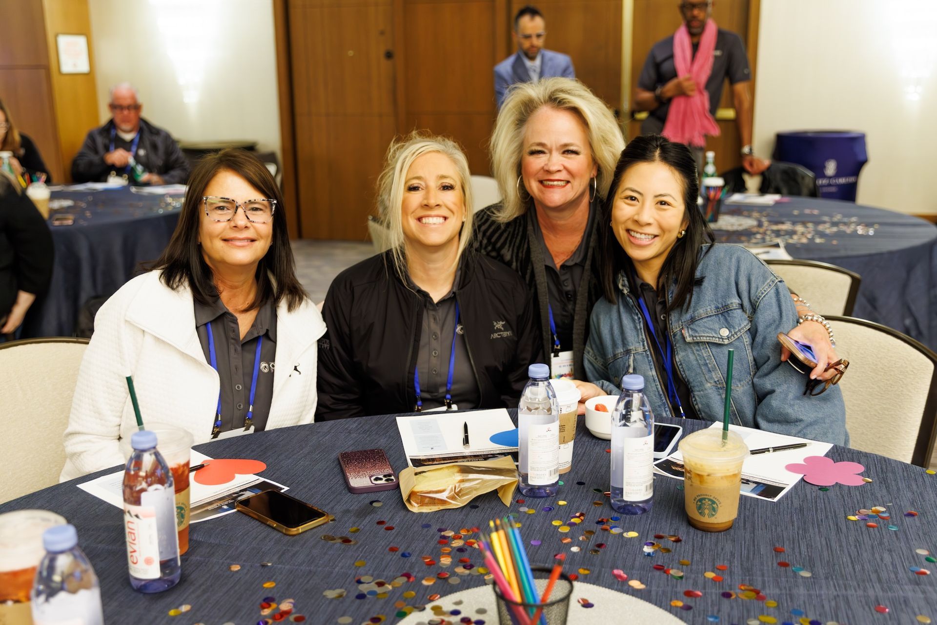 Four people smiling around a round conference table covered in blue cloth and confetti, with others visible in the room.