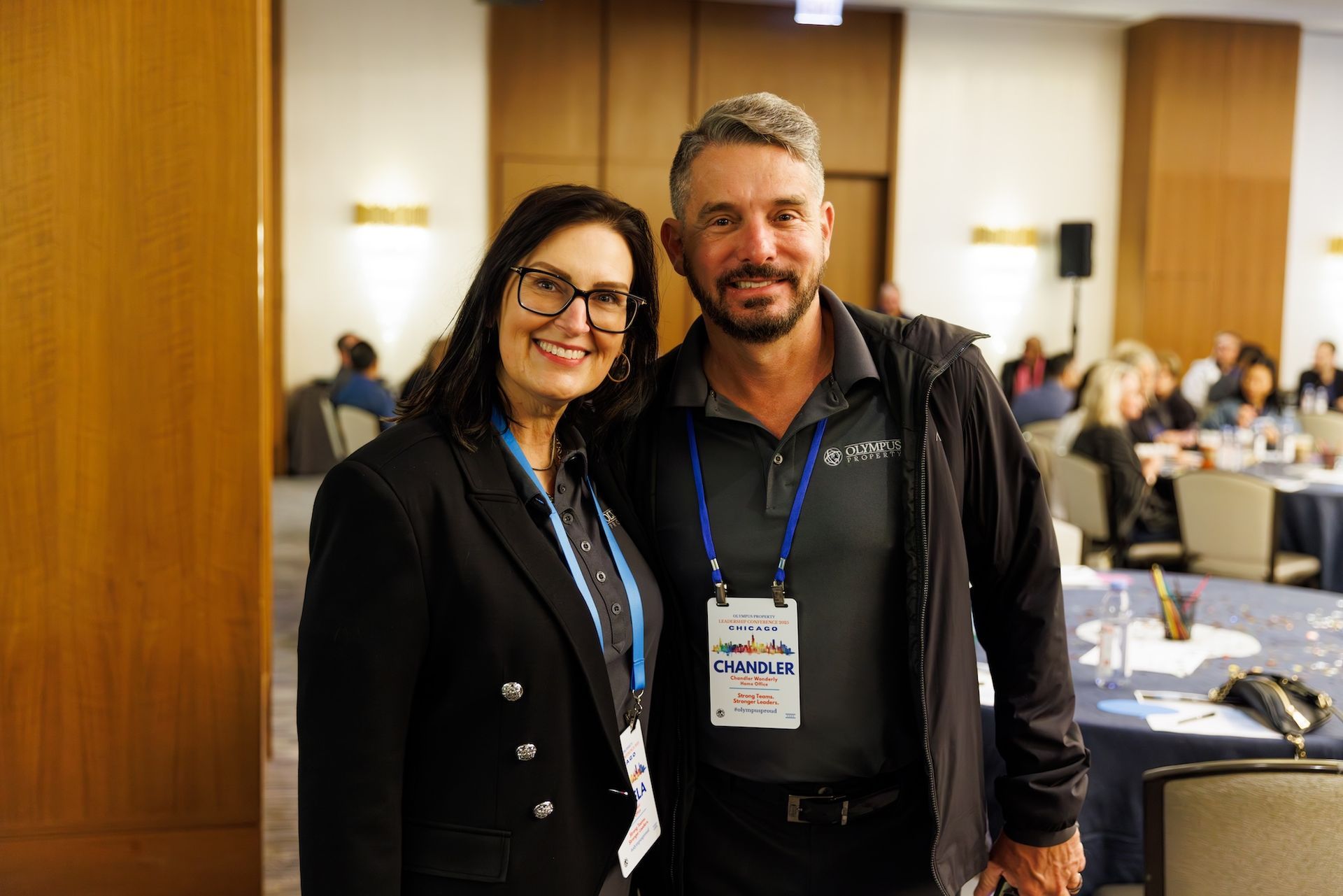 Two people standing in a conference room smiling at the camera, wearing event badges and business-casual attire.