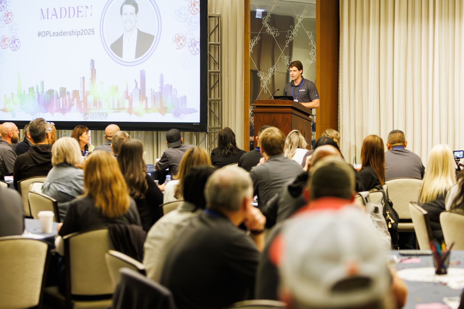 A speaker stands at a podium addressing an audience in a conference room with a large screen displaying 