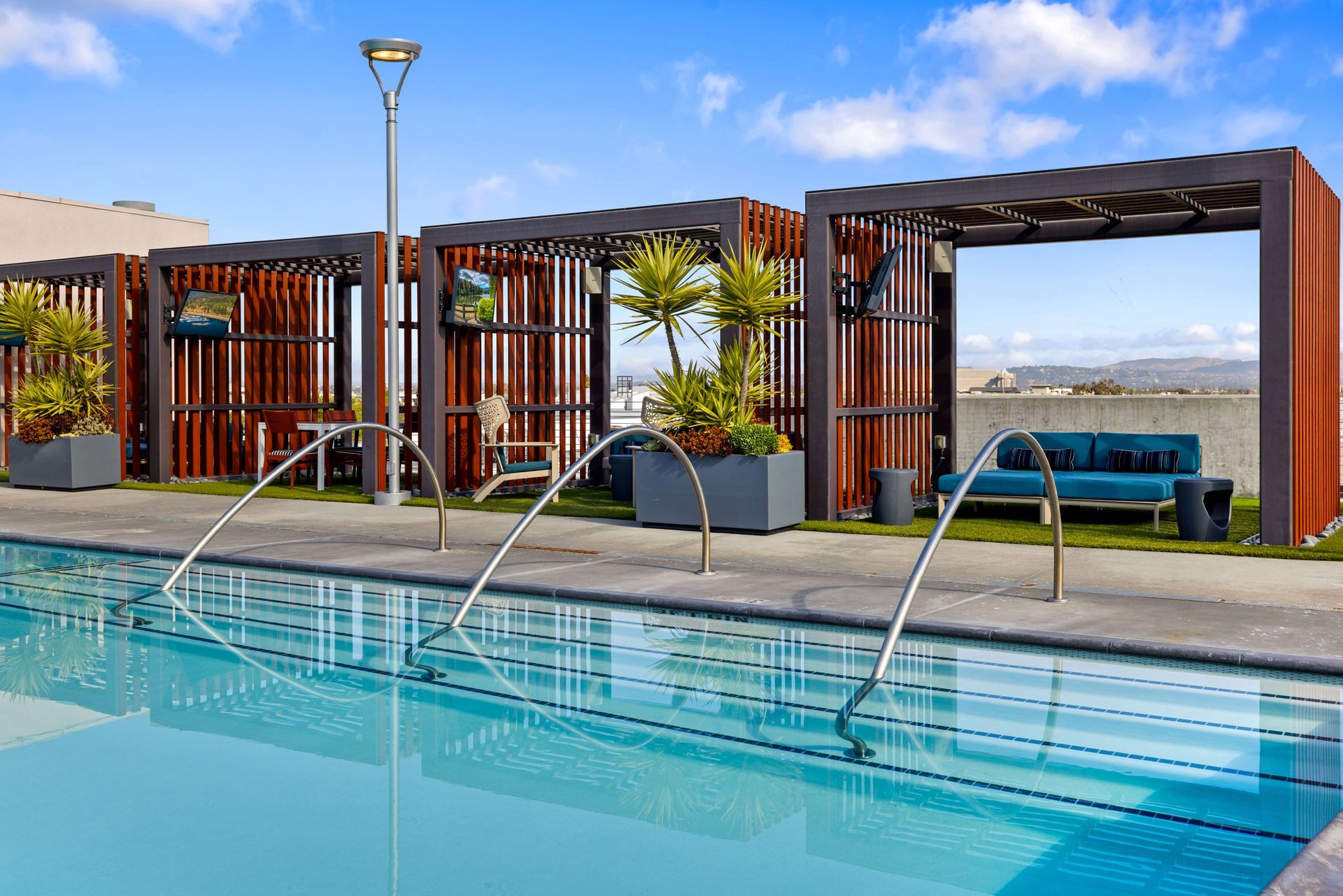 A swimming pool with metal handrails leading to a poolside lounge area featuring wooden cabanas, furniture, and plants.