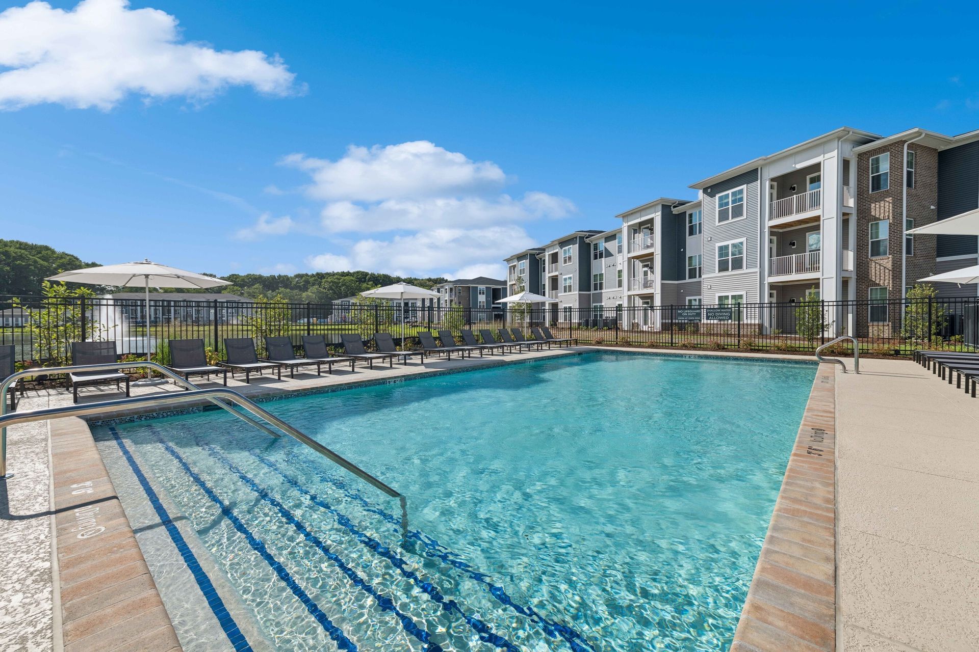 A swimming pool in front of a modern apartment complex under a bright blue sky with white clouds.