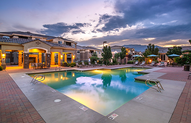 A swimming pool at a residential complex at sunset, with a multi-story building and patio seating in the background.