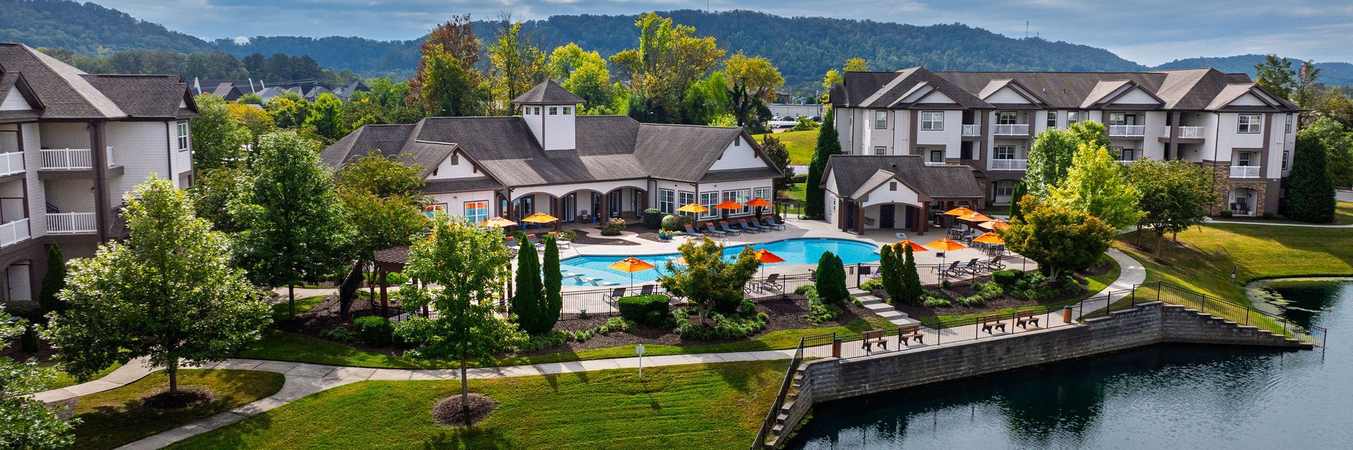 An aerial view of a multi-story apartment complex with a central swimming pool, surrounded by trees and a pond.