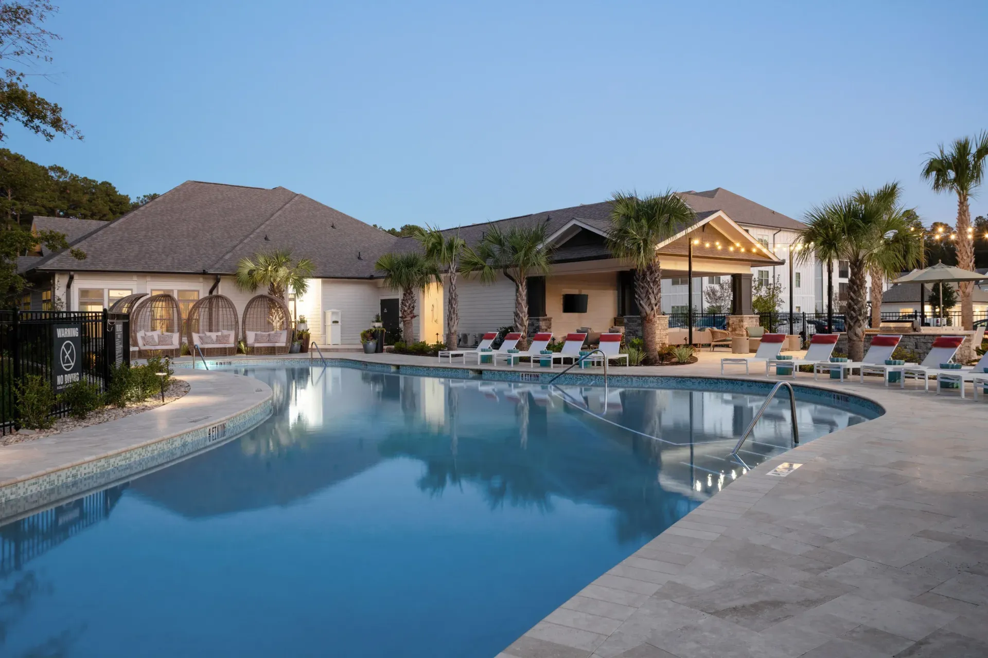 An outdoor swimming pool at a resort during dusk, surrounded by lounge chairs, palm trees, and a clubhouse.