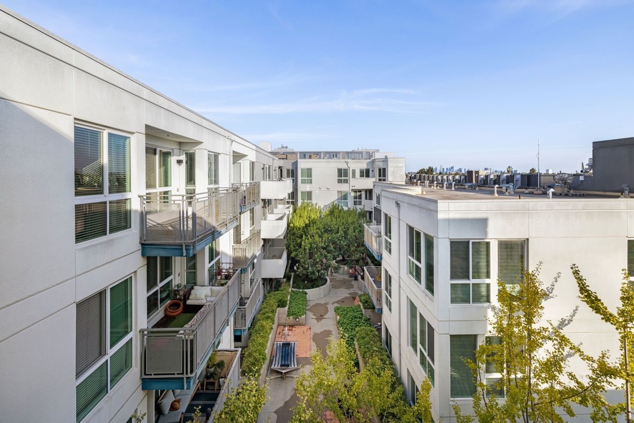 Two modern, white multi-story apartment buildings face each other, separated by a central courtyard with trees.