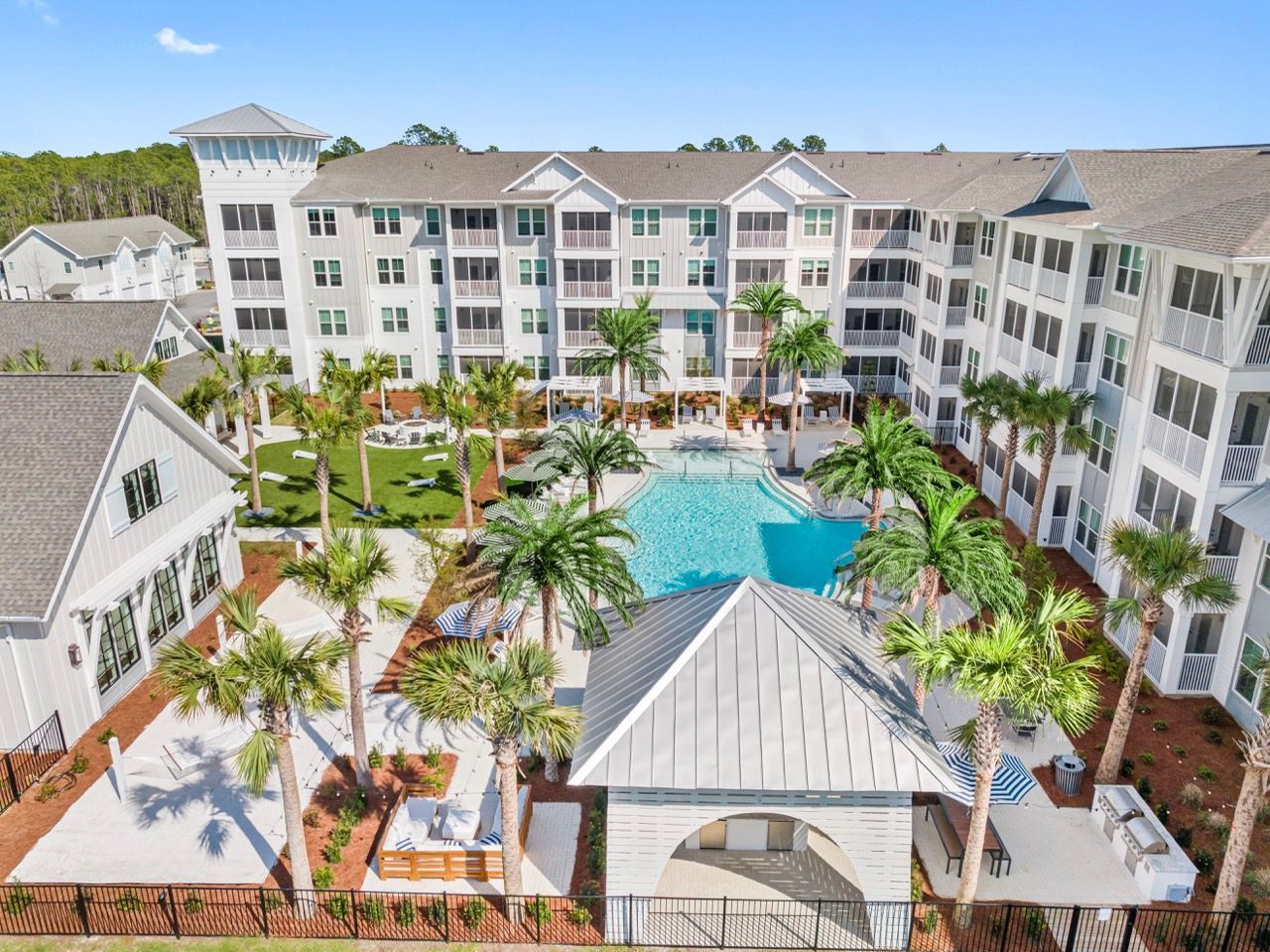 Aerial view of a white multi-story apartment complex with a central swimming pool, palm trees, and lounge areas.
