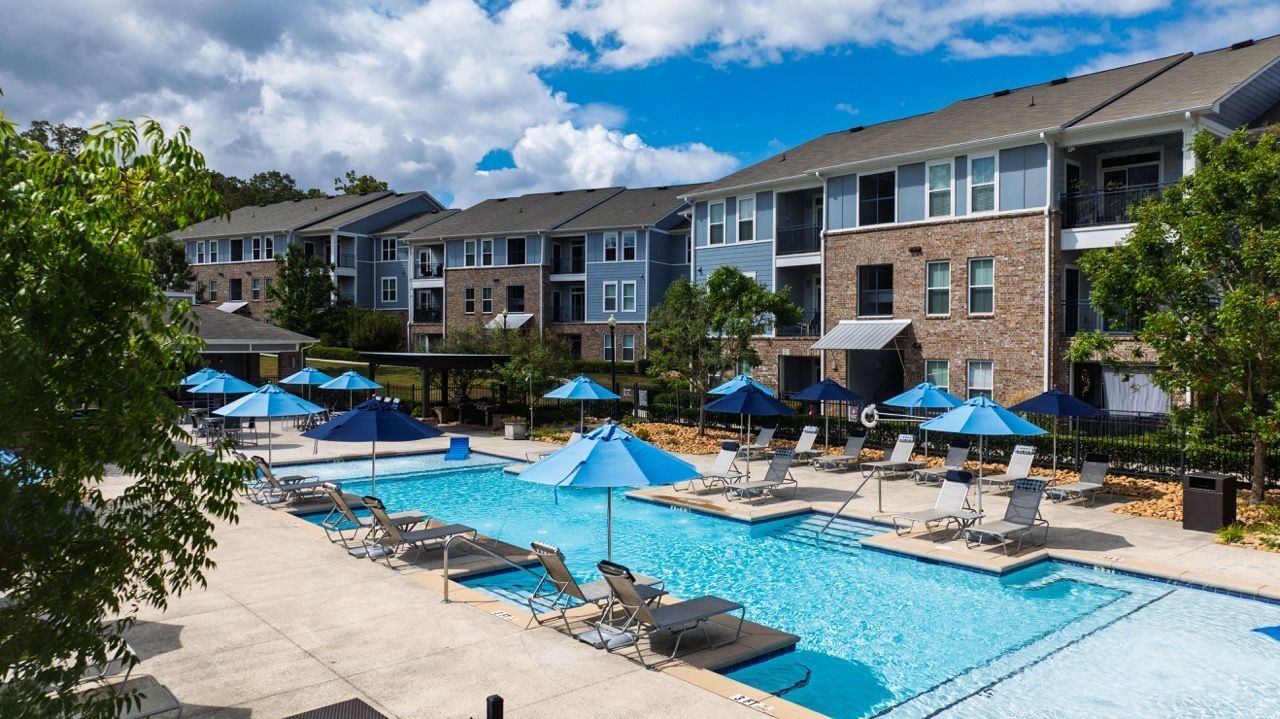 A swimming pool with blue umbrellas and lounge chairs in front of a multi-story apartment building under a cloudy sky.