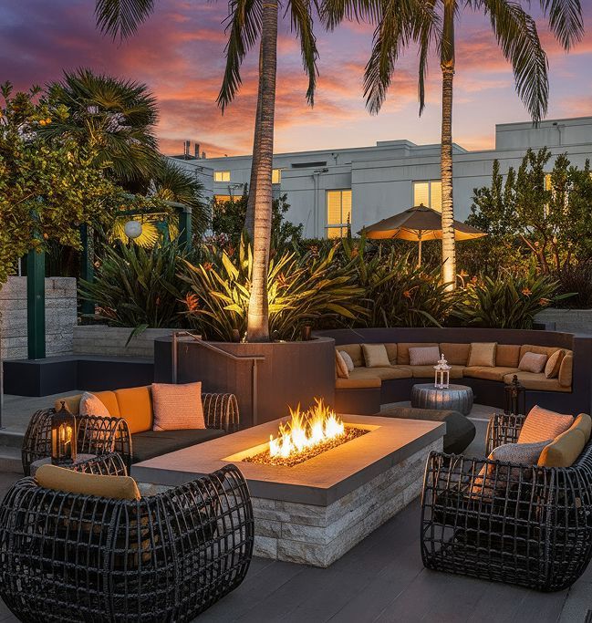 An outdoor patio at sunset with a central stone fire pit, modern wicker lounge chairs, and palm trees against a building.