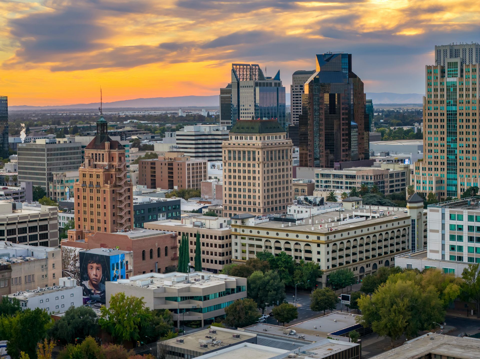 A cityscape of downtown Sacramento at sunset, featuring prominent tall buildings and golden light over the horizon.
