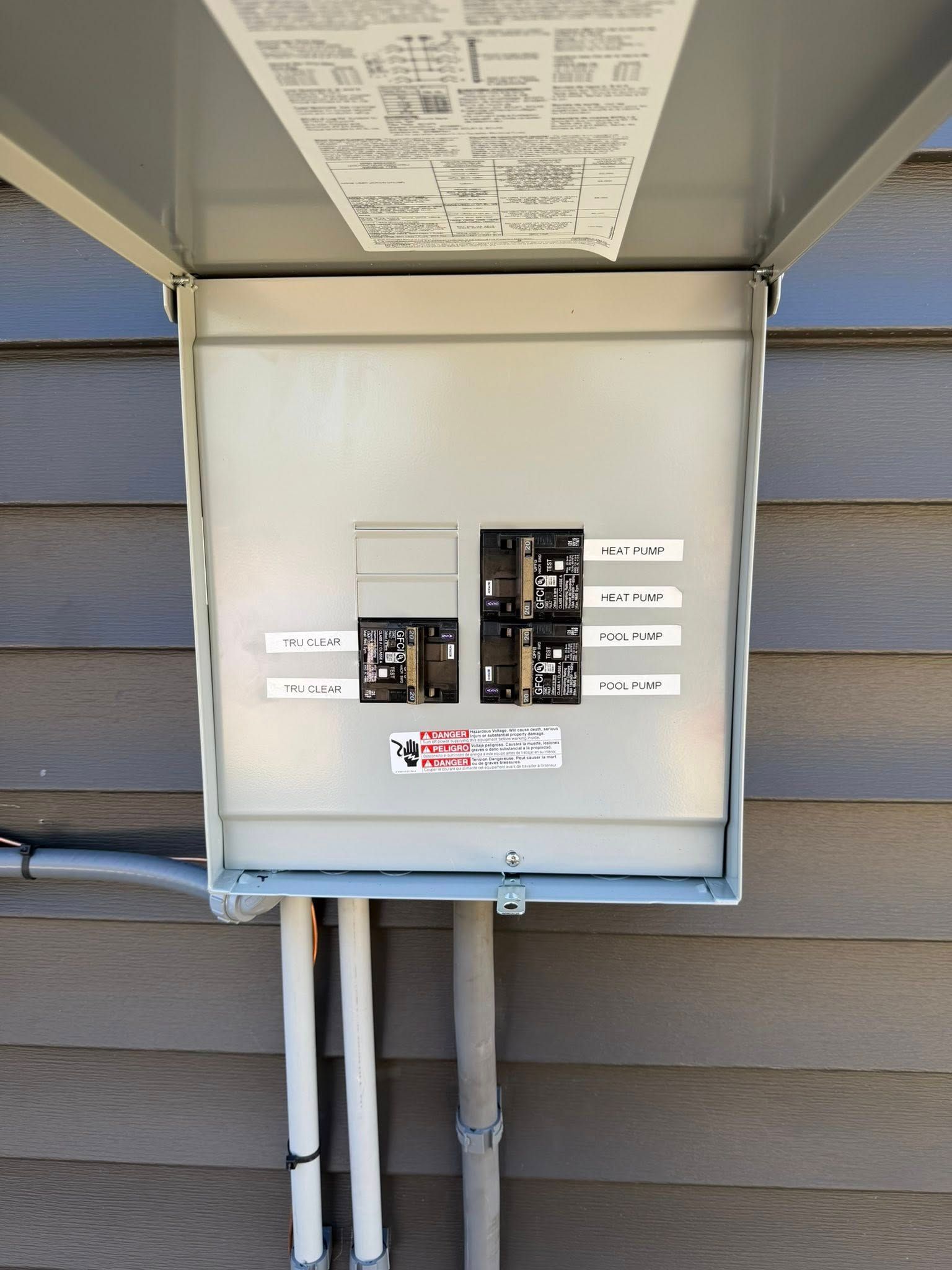 Outdoor electrical service panel on house siding with open breakers and conduit pipes below.