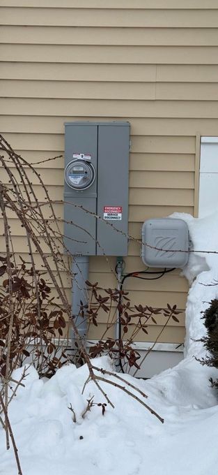 Gray utility meter boxes on beige siding beside a snowy yard and leafless shrubs