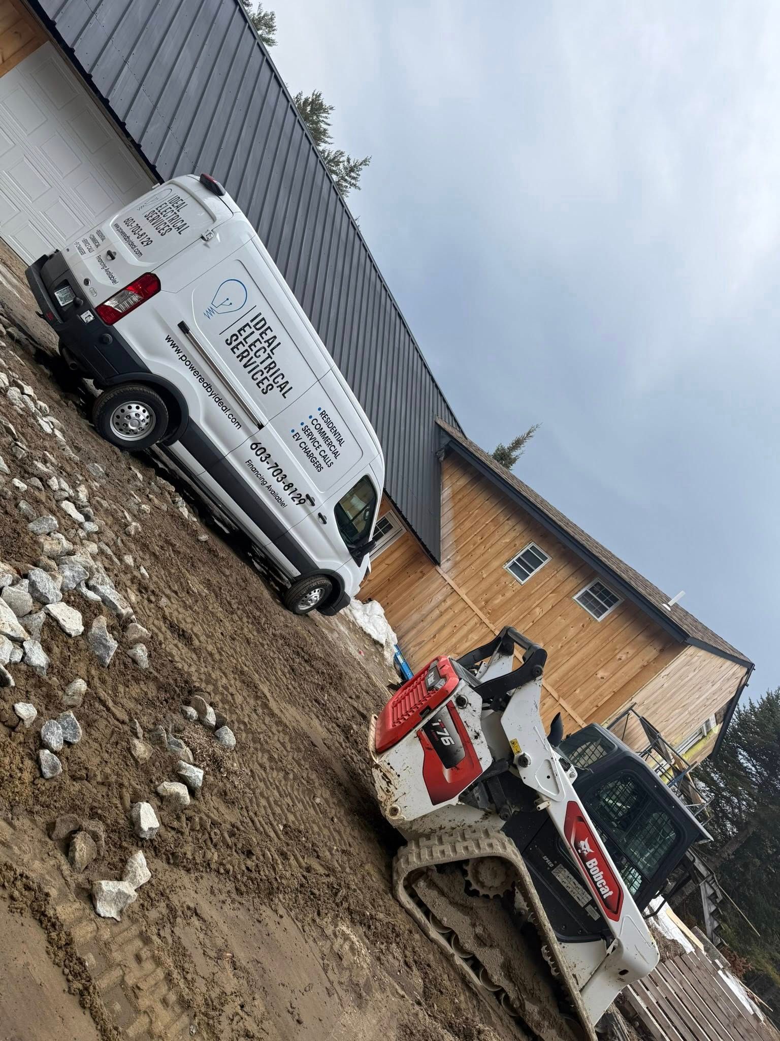 White work van and skid steer on muddy construction site beside a wooden building.