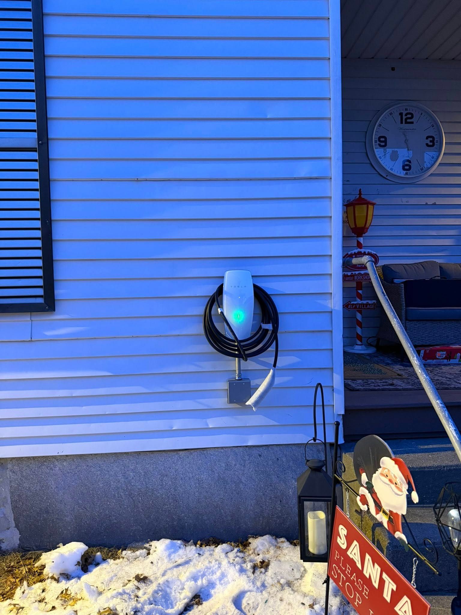 Blue-sided house exterior with wall-mounted EV charger, snow on the ground, and a clock on the wall.