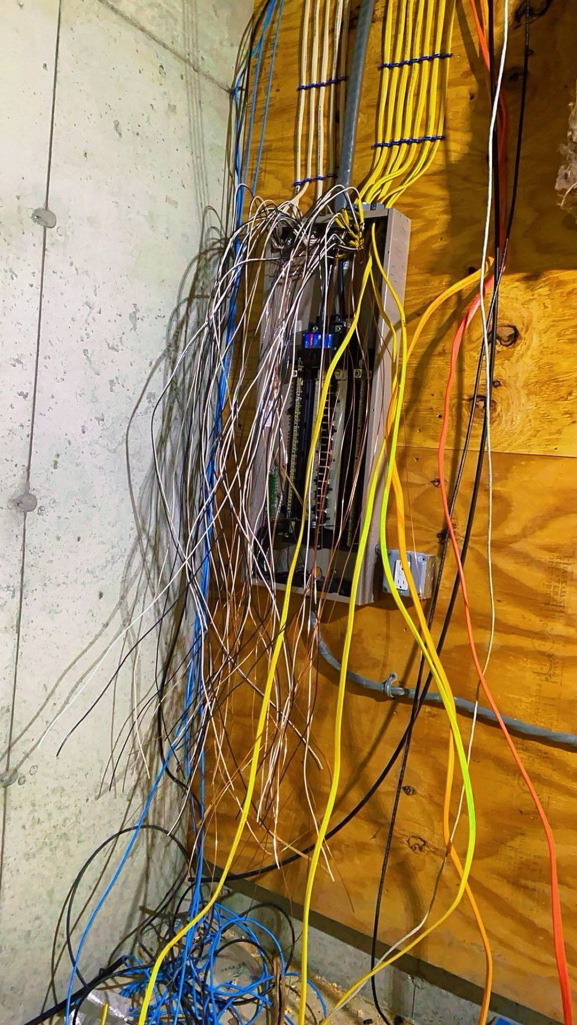 Dense bundle of tangled electrical wires hanging against a yellow wall in a narrow utility space