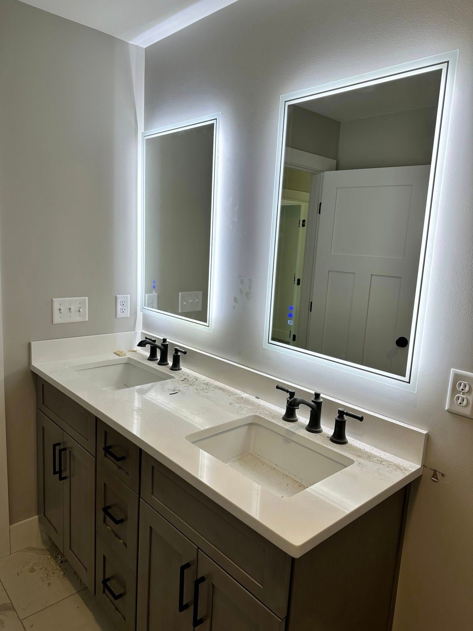 Bathroom vanity with double sinks, large lit mirrors, and dark cabinets against light walls.