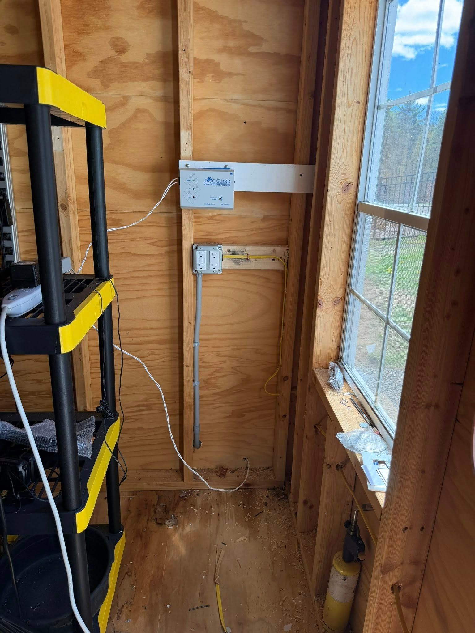 Small shed interior with electrical boxes, exposed wiring, shelving, and a window on the right.