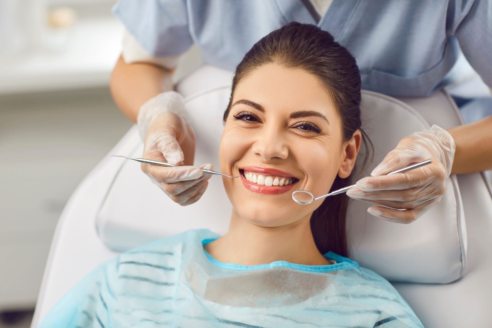 A dentist in a white coat and blue gloves holds a dental tool while smiling at a patient in a dental chair.