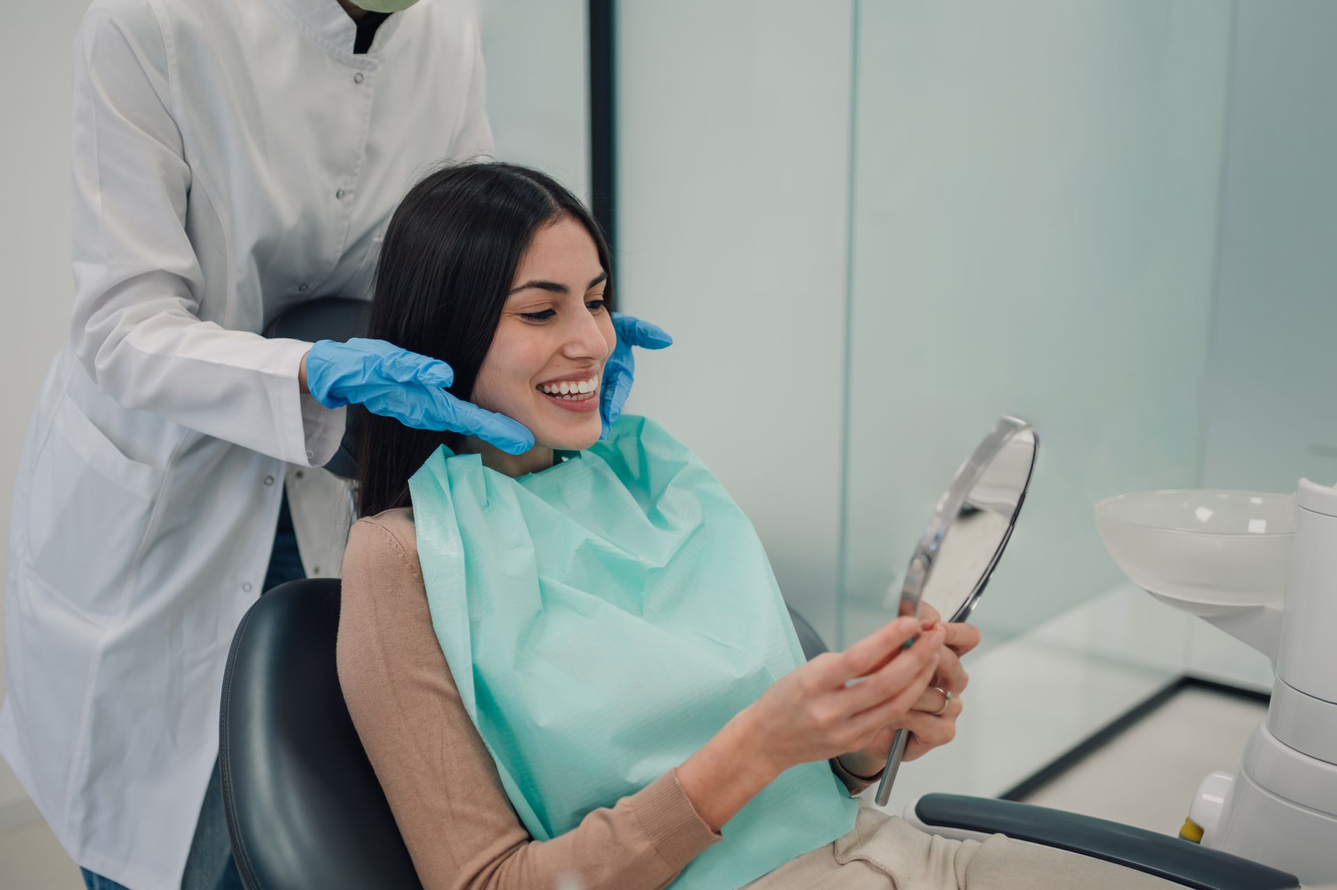 Dentist adjusting a patient’s mouth while she smiles and checks a handheld mirror in a clinic chair