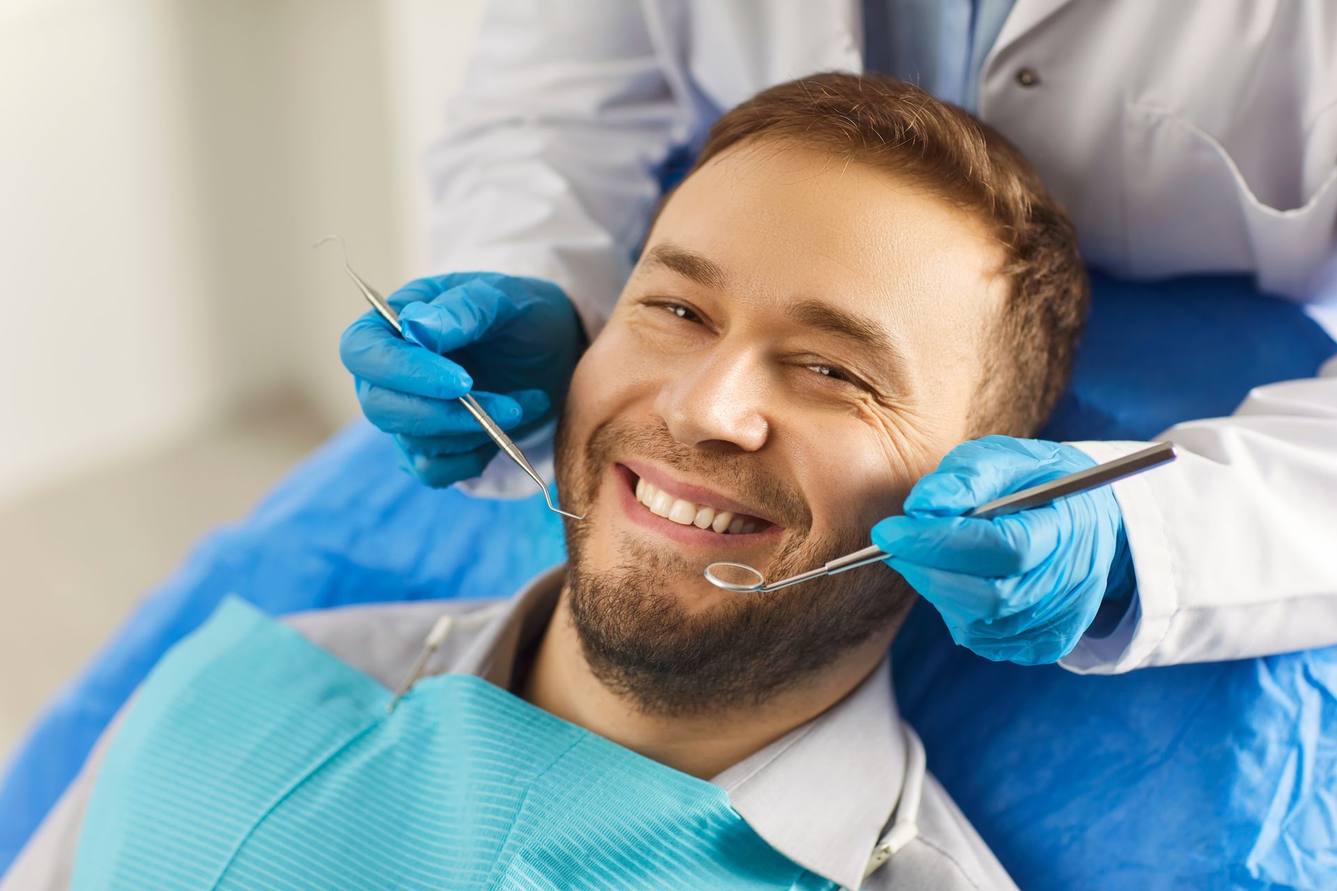 A person smiles while sitting in a dental chair as a professional holds a dental mirror and explorer near their mouth.