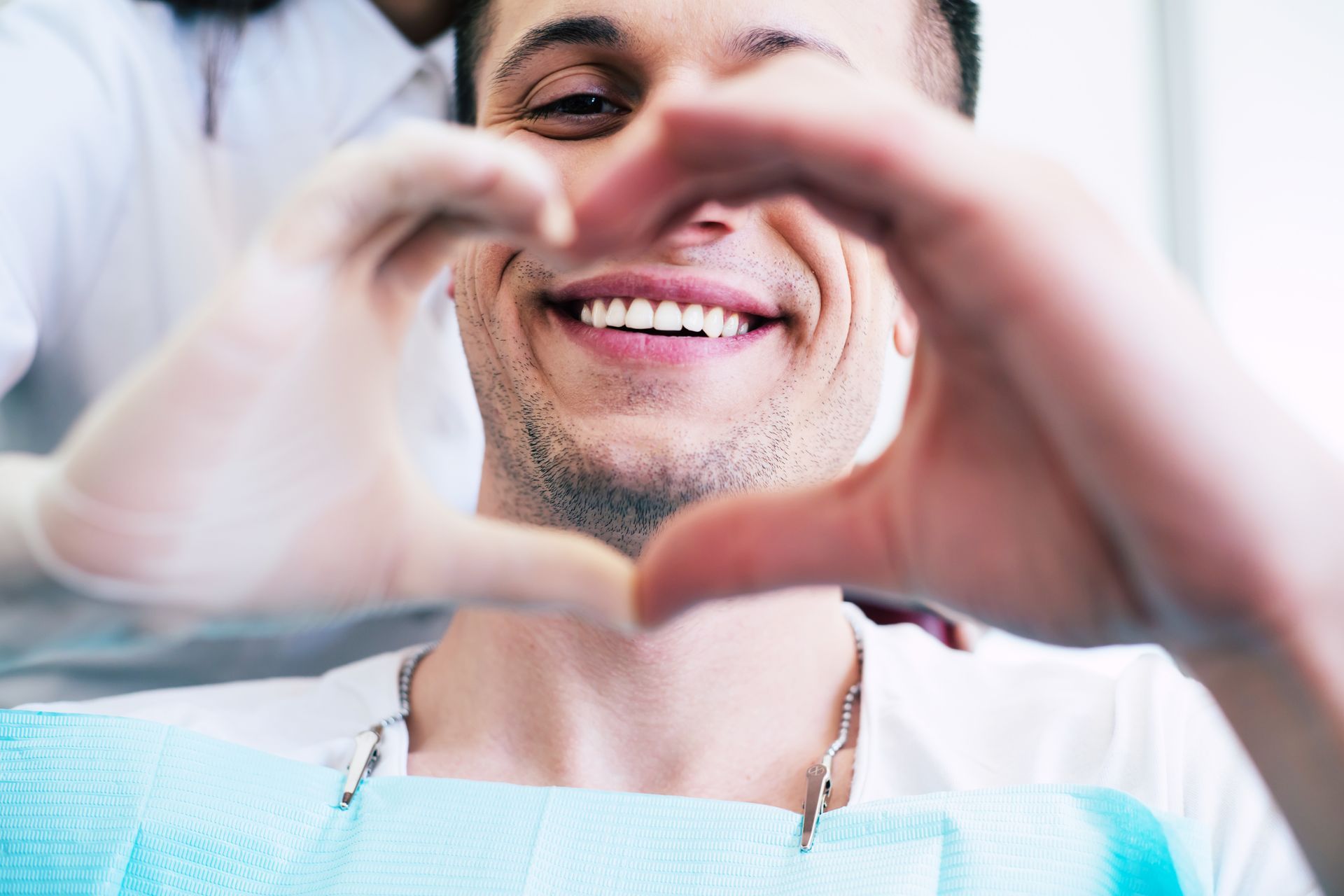 A person in a dental chair smiles while hands form a heart shape in front of their face.