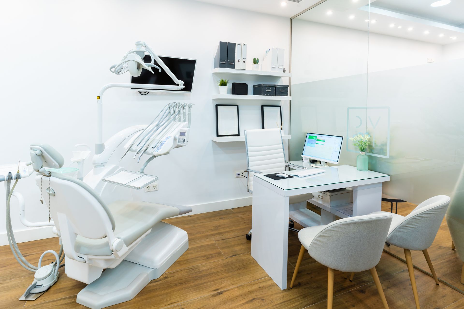Modern dental office featuring a patient chair, a desk with a computer, and chairs with white furniture and wood flooring.