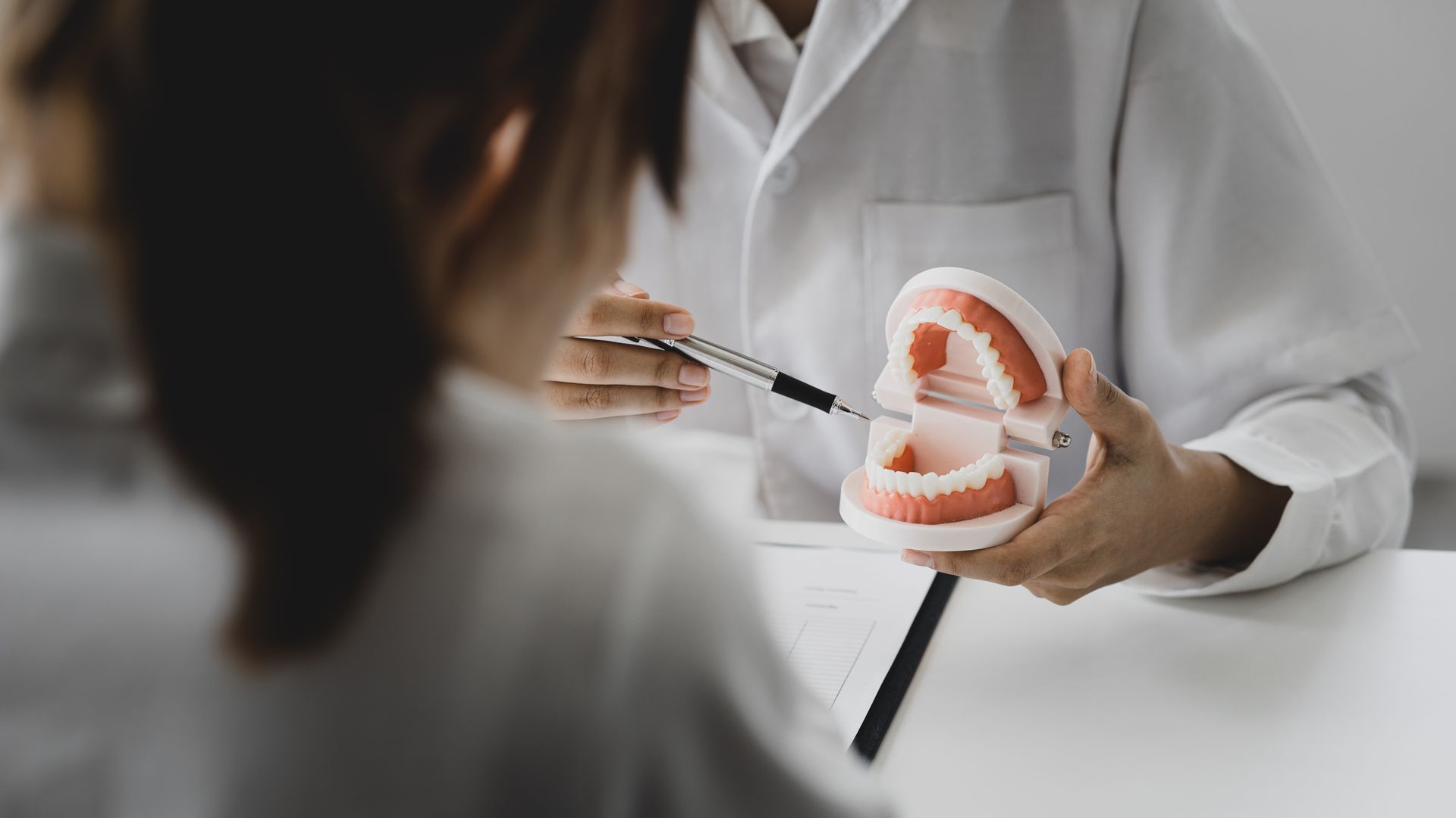 A person in a white lab coat uses a metal tool to explain a dental model to a patient in a medical office setting.