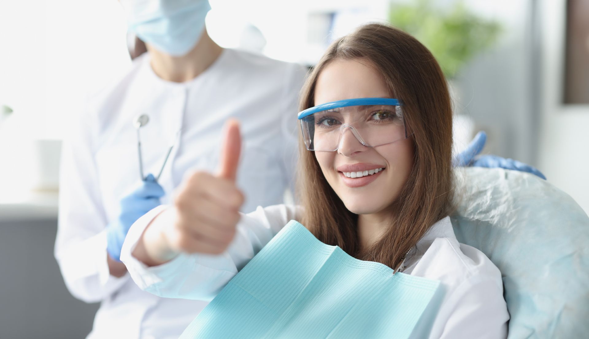 A smiling patient in protective eyewear gives a thumbs up in a dental office, with a masked clinician standing behind.
